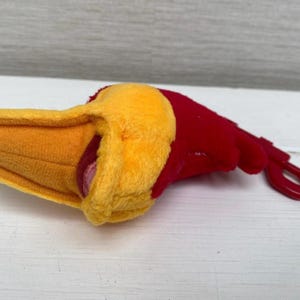 May include: A plush toy shaped like a bird's head, featuring a yellow beak and a red head. The toy has a red plastic loop attached. The toy is lying on a white surface.