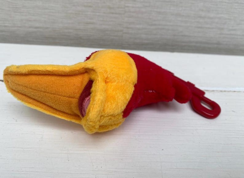 May include: A plush toy shaped like a bird's head, featuring a yellow beak and a red head. The toy has a red plastic loop attached. The toy is lying on a white surface.