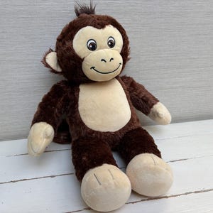 Puede incluir: Un peluche de mono con pelaje marrón oscuro en la cabeza, brazos y piernas, con un rostro, pecho, manos y pies en color beige claro. El mono tiene ojos negros bordados, una nariz cosida y una sonrisa curva en su rostro.