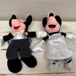 May include: Two plush Mickey and Minnie Mouse dolls dressed for a wedding. Mickey wears a black tuxedo with a white bow tie and Minnie wears a white wedding dress and veil. Both have black ears, pink faces, and red mouths.