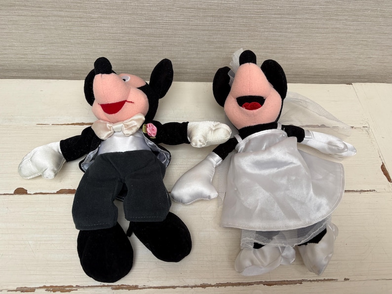 May include: Two plush Mickey and Minnie Mouse dolls dressed for a wedding. Mickey wears a black tuxedo with a white bow tie and Minnie wears a white wedding dress and veil. Both have black ears, pink faces, and red mouths.