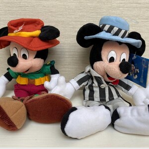 May include: Two Mickey Mouse plush toys. One wears a red hat with a yellow band, a green shirt, and maroon trousers. The other Mickey wears a blue hat with black and white stripes and a black and white striped jacket. Both have white gloves.