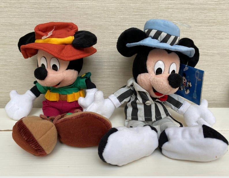 May include: Two Mickey Mouse plush toys. One wears a red hat with a yellow band, a green shirt, and maroon trousers. The other Mickey wears a blue hat with black and white stripes and a black and white striped jacket. Both have white gloves.
