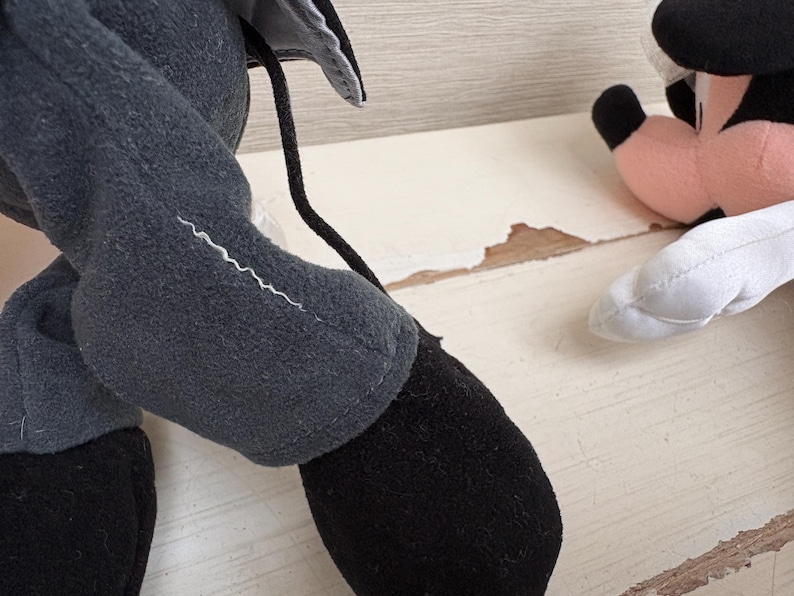 May include: Close-up of a plush toy's leg and foot, featuring a black shoe and gray pants. A white zigzag stitch detail is visible on the leg. Another plush toy is partially visible in the background.