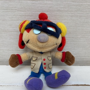 May include: A plush toy character featuring a tan face, a red hat with yellow ear flaps, and a black mask over the eyes. The figure wears a beige vest with red buttons over a blue shirt, brown gloves, and purple shoes with yellow accents.