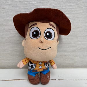 May include: A plush toy of the character Woody from the movie Toy Story. The toy has a brown cowboy hat, a tan face, and a yellow shirt with a cow print vest. It is wearing blue jeans and brown boots.