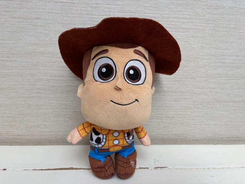 May include: A plush toy of the character Woody from the movie Toy Story. The toy has a brown cowboy hat, a tan face, and a yellow shirt with a cow print vest. It is wearing blue jeans and brown boots.