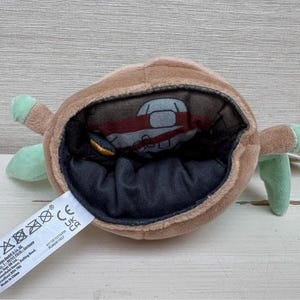 May include: A brown plush toy with a large opening, revealing a smaller figure inside. The toy has green appendages and a black interior. A white tag with text is attached.