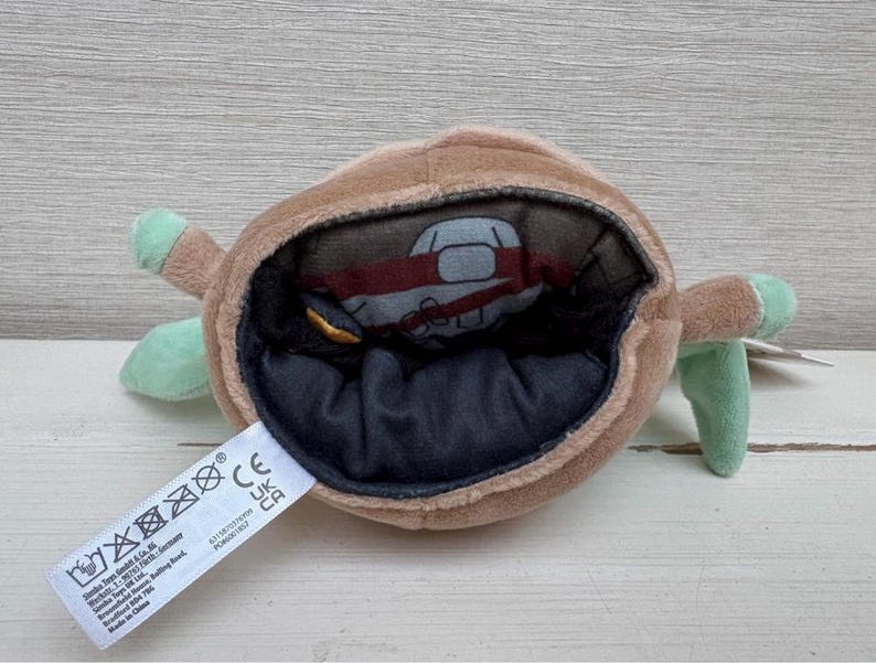 May include: A brown plush toy with a large opening, revealing a smaller figure inside. The toy has green appendages and a black interior. A white tag with text is attached.