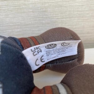 May include: Close-up of a plush toy with a white tag. The toy is primarily brown and dark blue, with red and white accents. The tag has the letters "UK CA" and the CE mark.