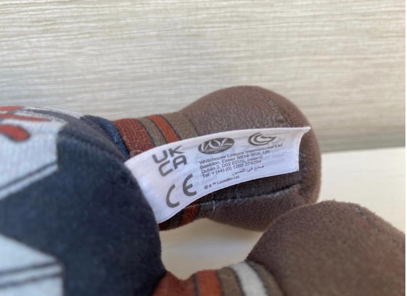 May include: Close-up of a plush toy with a white tag. The toy is primarily brown and dark blue, with red and white accents. The tag has the letters "UK CA" and the CE mark.