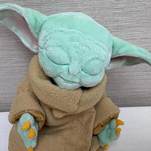 May include: A soft toy of a character with large, pointed ears, a light green face, and closed eyes. The character wears a tan coat with light green hands and yellow accents. The toy is a cuddly item.