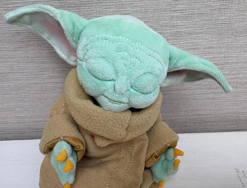 May include: A soft toy of a character with large, pointed ears, a light green face, and closed eyes. The character wears a tan coat with light green hands and yellow accents. The toy is a cuddly item.