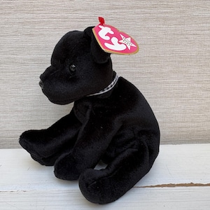 Puede incluir: Un juguete de peluche negro con forma de perro, con un collar blanco y negro y una etiqueta roja. El perro está sentado con las patas extendidas. La etiqueta es rosa y blanca con una estrella y las palabras "Beanie Baby".
