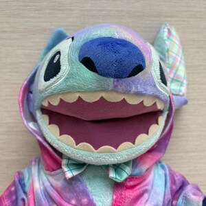 May include: A plush Stitch character in a colorful tie-dye outfit. The toy has large black eyes, a blue nose, and a wide open mouth with white teeth. The outfit includes a hood and a bow tie, with a mix of pink, purple, and blue hues.
