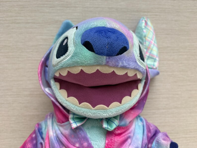 May include: A plush Stitch character in a colorful tie-dye outfit. The toy has large black eyes, a blue nose, and a wide open mouth with white teeth. The outfit includes a hood and a bow tie, with a mix of pink, purple, and blue hues.