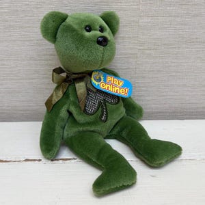 May include: A green plush teddy bear with a black nose and a dark green bow. The bear has a shamrock on its chest and a tag that says "Play Online!"