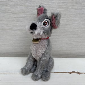 May include: A plush dog toy with grey fur, a lighter grey chest, and pink inner ears. The dog has a black nose, brown eyes, and a red collar with a small gold-colored tag. The toy is in a seated position with a shaggy texture around the muzzle.