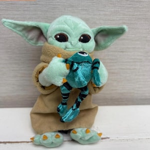 May include: A plush toy of a green alien character with large ears, holding a teal frog. The alien is wearing a tan robe and has large black eyes. The frog is teal with black stripes.
