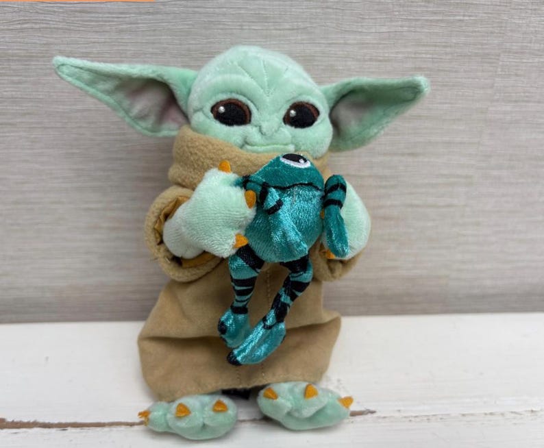 May include: A plush toy of a green alien character with large ears, holding a teal frog. The alien is wearing a tan robe and has large black eyes. The frog is teal with black stripes.