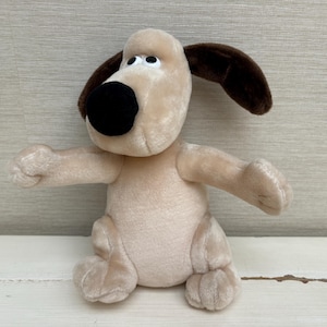 Puede incluir: Un peluche de perro con pelaje beige claro, una nariz grande y redondeada de color negro y orejas de color marrón oscuro. El animal de peluche está sentado con los brazos extendidos hacia adelante y tiene ojos blancos con pupilas negras mirando hacia arriba.