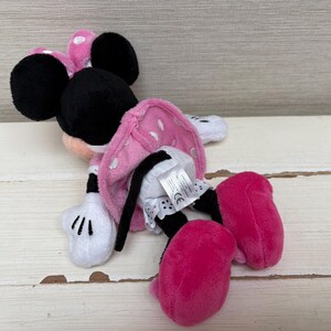 May include: A plush Minnie Mouse toy, featuring a pink polka-dot dress, large black ears, and bright pink shoes. The toy has white gloves and is lying on a light-colored wooden surface. The toy is a classic Disney character.