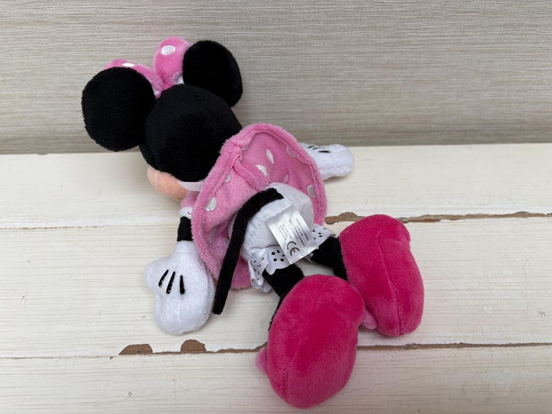 May include: A plush Minnie Mouse toy, featuring a pink polka-dot dress, large black ears, and bright pink shoes. The toy has white gloves and is lying on a light-colored wooden surface. The toy is a classic Disney character.