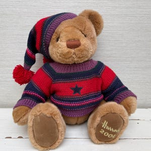 May include: A plush teddy bear wearing a striped knit cap with a red pom-pom and a matching sweater. The sweater has a star and the bear's foot has "Harrods 2004" embroidered on it. The bear is brown with a brown nose and paws.