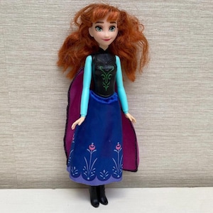 May include: A doll of a character with red hair, wearing a black top with green accents, a blue skirt with floral designs, and a purple cape. The doll has black boots and is standing against a light brown background.
