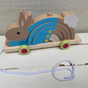 May include: A wooden pull-along toy shaped like a bunny rabbit with a rainbow. The bunny is tan with blue accents and a white cloud. The rainbow is blue, tan, and white. The toy has green wheels with pink centers and a white pull string.