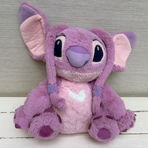 May include: A plush toy of the character Angel from the Lilo & Stitch franchise. The toy is primarily lavender with pink accents on the ears, chest, and feet. It has large black eyes and a purple nose. The toy is sitting on a white wooden surface.