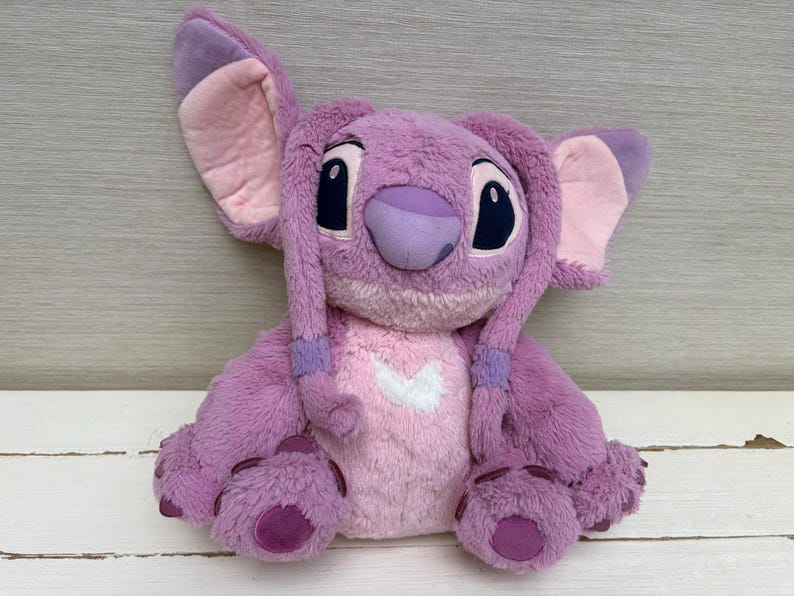 May include: A plush toy of the character Angel from the Lilo & Stitch franchise. The toy is primarily lavender with pink accents on the ears, chest, and feet. It has large black eyes and a purple nose. The toy is sitting on a white wooden surface.