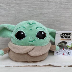 May include: A Grogu plush toy from The Mandalorian series. The toy has large black eyes, green skin, and a tan robe. A Star Wars Mandalorian tag is attached.