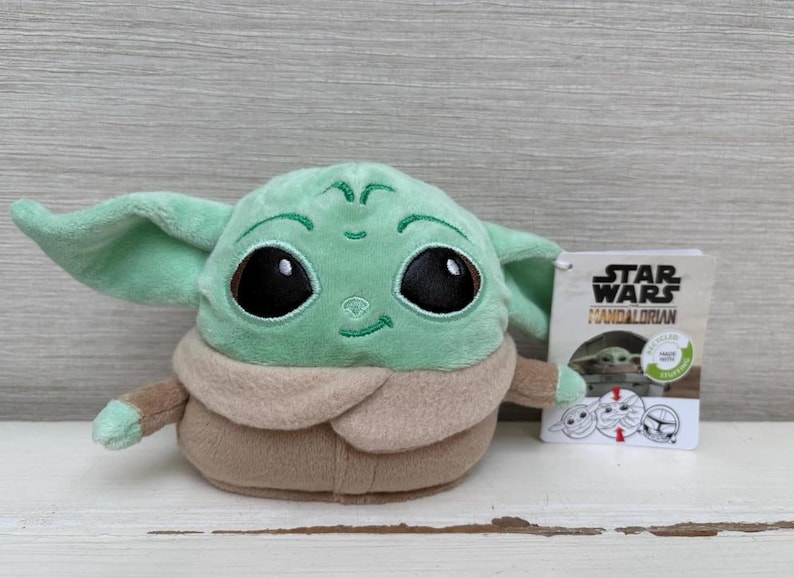 May include: A Grogu plush toy from The Mandalorian series. The toy has large black eyes, green skin, and a tan robe. A Star Wars Mandalorian tag is attached.