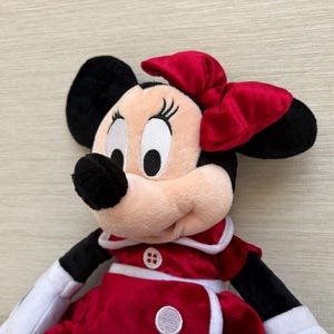 May include: A Minnie Mouse soft toy featuring a red bow, black ears, and a red and white dress. The doll has a smiling expression and is made of soft, plush materials. A classic Disney character.