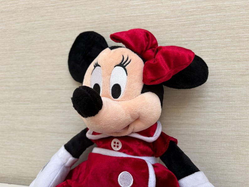 May include: A Minnie Mouse soft toy featuring a red bow, black ears, and a red and white dress. The doll has a smiling expression and is made of soft, plush materials. A classic Disney character.
