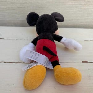 K&ouml;nnte beinhalten: Ein kleines Mickey Mouse Pl&uuml;schtier. Das Spielzeug hat einen schwarzen Kopf und Ohren, wei&szlig;e Handschuhe, rote Shorts und gelbe Schuhe. Ein wei&szlig;es Etikett ist am Spielzeug befestigt.