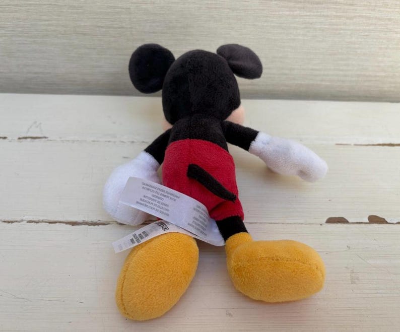K&ouml;nnte beinhalten: Ein kleines Mickey Mouse Pl&uuml;schtier. Das Spielzeug hat einen schwarzen Kopf und Ohren, wei&szlig;e Handschuhe, rote Shorts und gelbe Schuhe. Ein wei&szlig;es Etikett ist am Spielzeug befestigt.