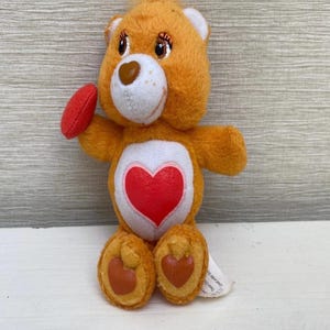 May include: An orange plush bear with a red heart on its chest and a small red heart in its paw. The bear has a white belly, brown eyes, and a brown nose. It is a vintage toy.
