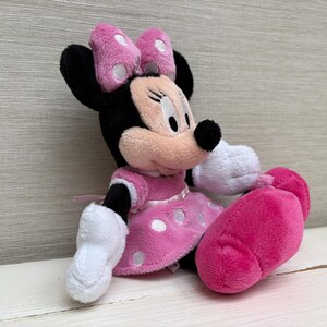 May include: A plush Minnie Mouse doll in a seated position. The doll features a black head and ears, a pink bow with white polka dots, and a pink dress with white polka dots. The doll's hands and feet are white and pink.