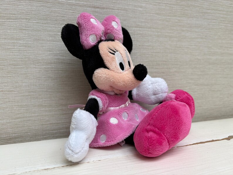 May include: A plush Minnie Mouse doll in a seated position. The doll features a black head and ears, a pink bow with white polka dots, and a pink dress with white polka dots. The doll's hands and feet are white and pink.