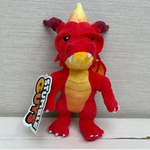 May include: A plush red dragon toy with yellow and orange accents. The dragon has white fangs, small horns, and a tag that reads "Stumble Guys". The toy is designed with a friendly, cartoonish appearance.