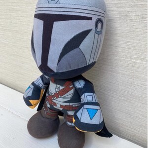 May include: A plush toy depicting a character in a gray and black helmet and armor. The toy has a rounded head, large feet, and a small cape. The character's helmet has a T-shaped visor and the toy is on a white surface.