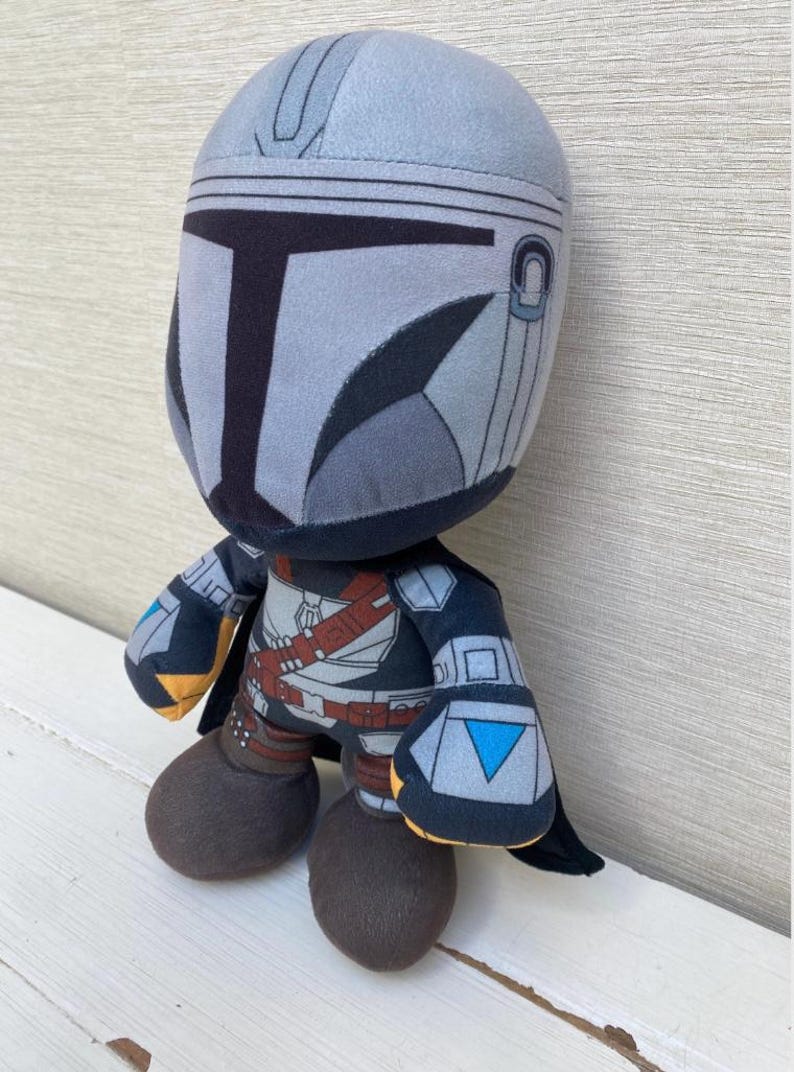 May include: A plush toy depicting a character in a gray and black helmet and armor. The toy has a rounded head, large feet, and a small cape. The character's helmet has a T-shaped visor and the toy is on a white surface.