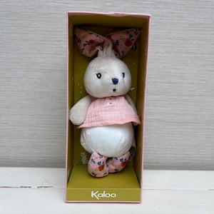 May include: A plush rabbit toy with white fur, wearing a light pink textured top. The rabbit has long ears with a pink floral pattern and matching floral fabric on its feet. It is packaged in an open-front box with the brand name Kaloo printed.