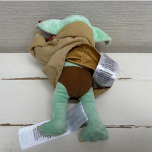 Pode incluir: Um brinquedo de pel&uacute;cia de um personagem da s&eacute;rie Star Wars, com cabe&ccedil;a e p&eacute;s verde claro, cal&ccedil;&otilde;es castanhos e uma t&uacute;nica bege. O brinquedo est&aacute; deitado de costas com uma etiqueta.