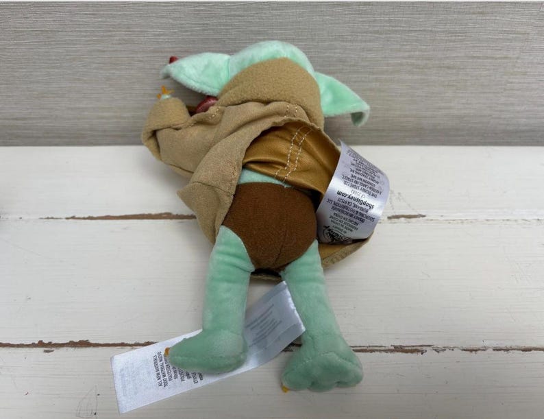 Pode incluir: Um brinquedo de pel&uacute;cia de um personagem da s&eacute;rie Star Wars, com cabe&ccedil;a e p&eacute;s verde claro, cal&ccedil;&otilde;es castanhos e uma t&uacute;nica bege. O brinquedo est&aacute; deitado de costas com uma etiqueta.