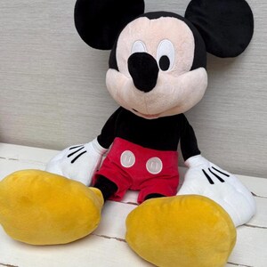 May include: A plush Mickey Mouse doll with large black ears, a black body, red shorts with white buttons, and yellow feet. The doll has white gloves with black lines and a smiling face.