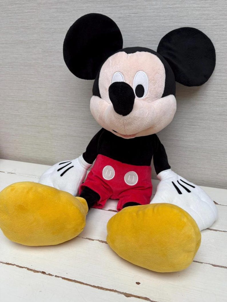May include: A plush Mickey Mouse doll with large black ears, a black body, red shorts with white buttons, and yellow feet. The doll has white gloves with black lines and a smiling face.