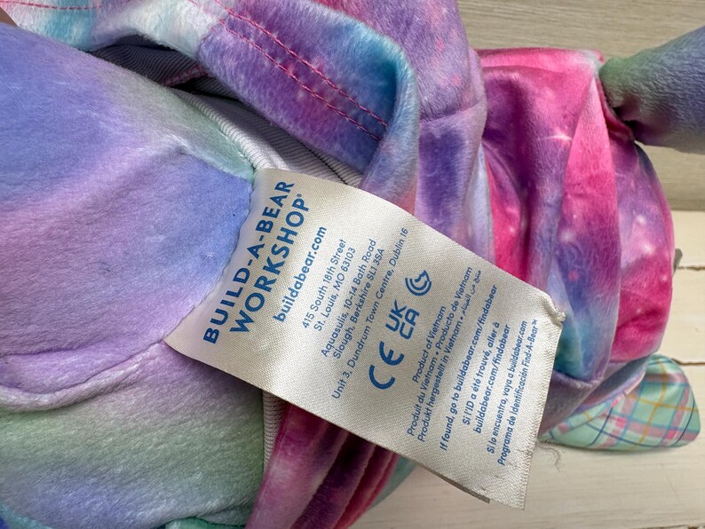 May include: A Build-A-Bear Workshop plush toy with a colorful tie-dye pattern in shades of pink, purple, blue, and green. The toy has a tag with the brand's logo and product information.